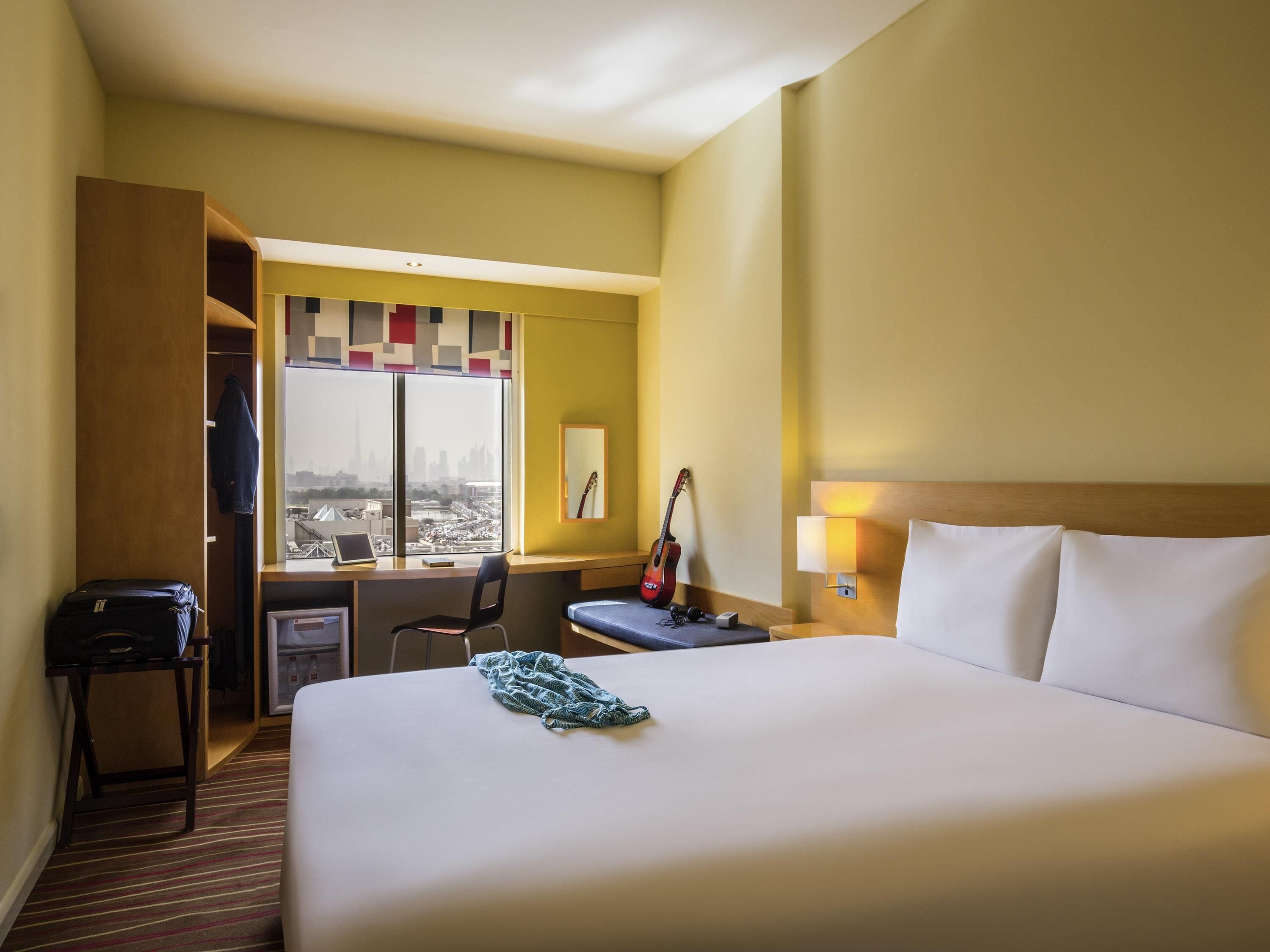 Une chambre d’hôtel avec un grand lit, un bureau avec un ordinateur portable, une guitare et une vue sur le paysage urbain par la fenêtre.