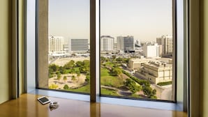 Quarto com uma grande janela com vista para a paisagem urbana, uma pequena mesa com um tablet e um par de óculos, e uma vista para um parque e edifícios.