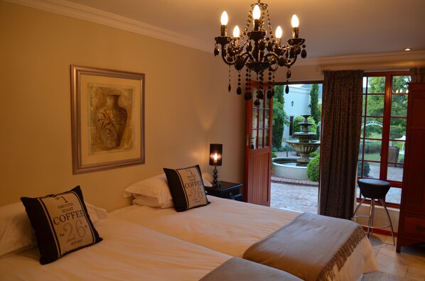A hotel room with two beds, a chandelier, a framed artwork, and a view of a garden through a window. - Cultivar Guest Lodge (Stellenbosch)
