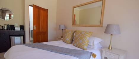 A neatly made bed with white bedding and patterned pillows, a bedside table with a lamp, and a mirror on the wall.