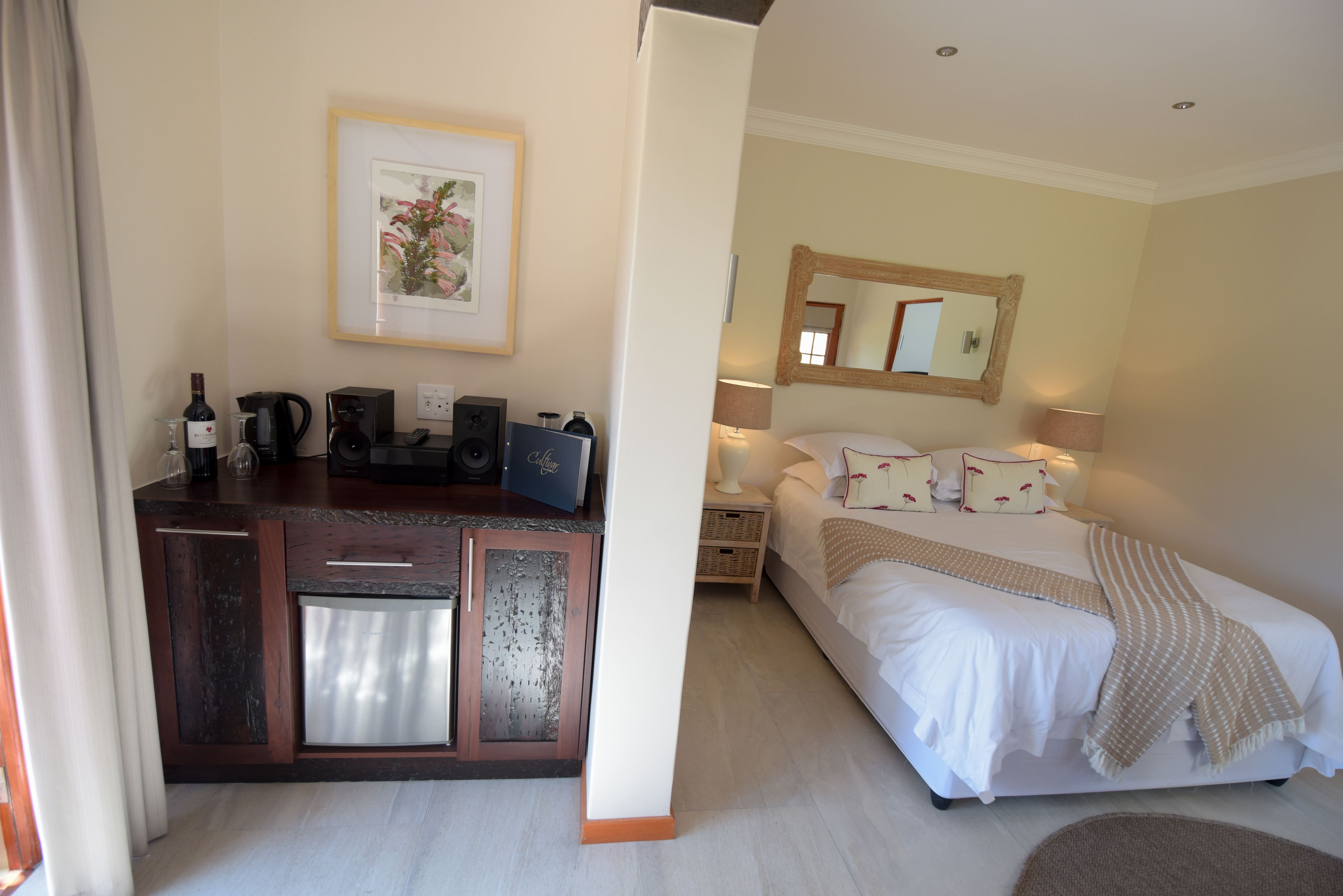 Habitación de hotel con cama, mesita de noche y un área de kitchenette con cafetera y botella de vino.