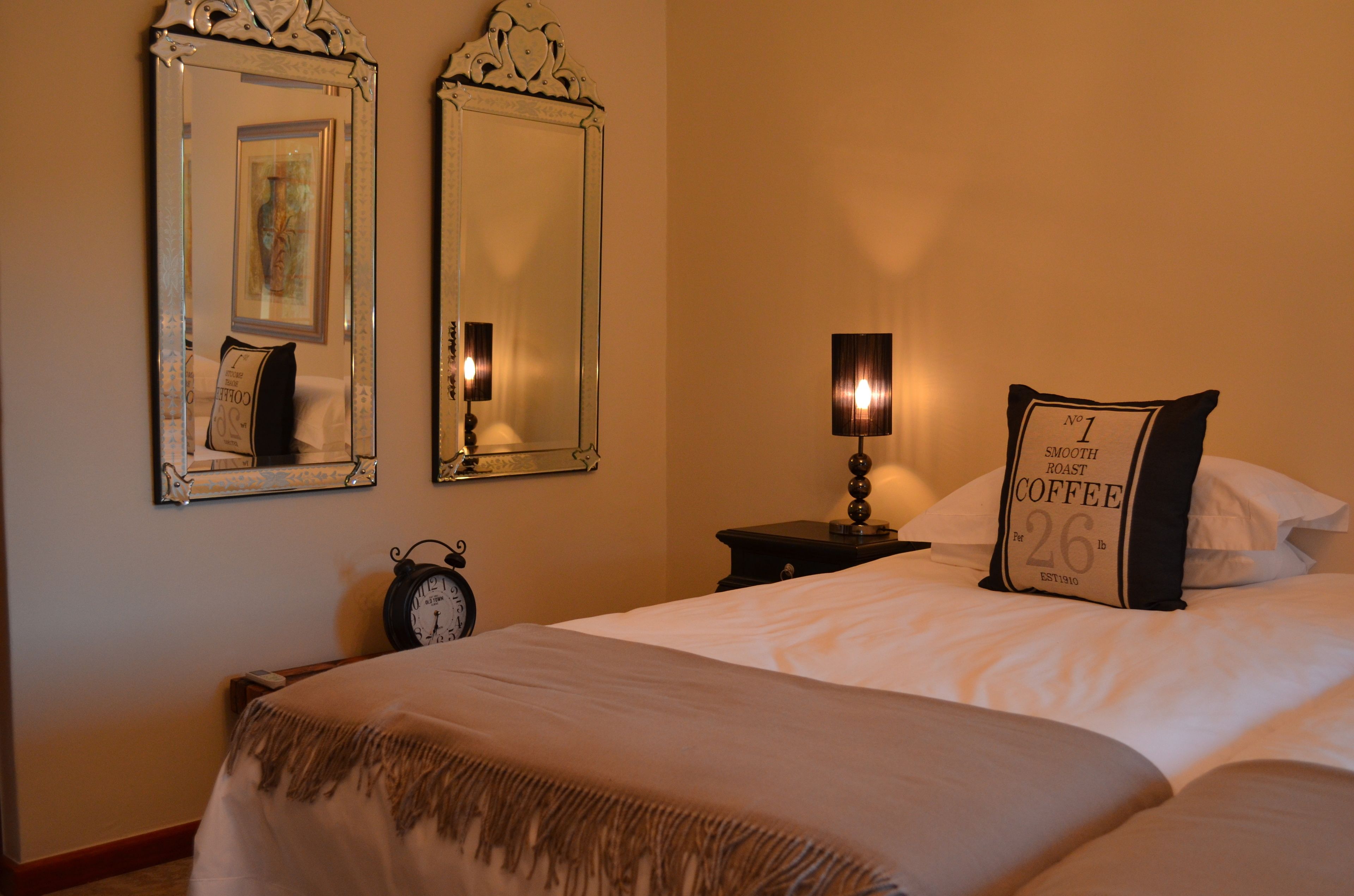 A neatly made bed with a pillow labeled "No 1 Smooth Roast Coffee 26 lb." Two wall mirrors and a small table with a lamp are also visible.