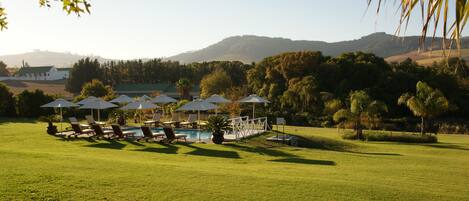 A pool area with lounge chairs and umbrellas, surrounded by a well-maintained lawn and trees, with a view of hills in the background.
