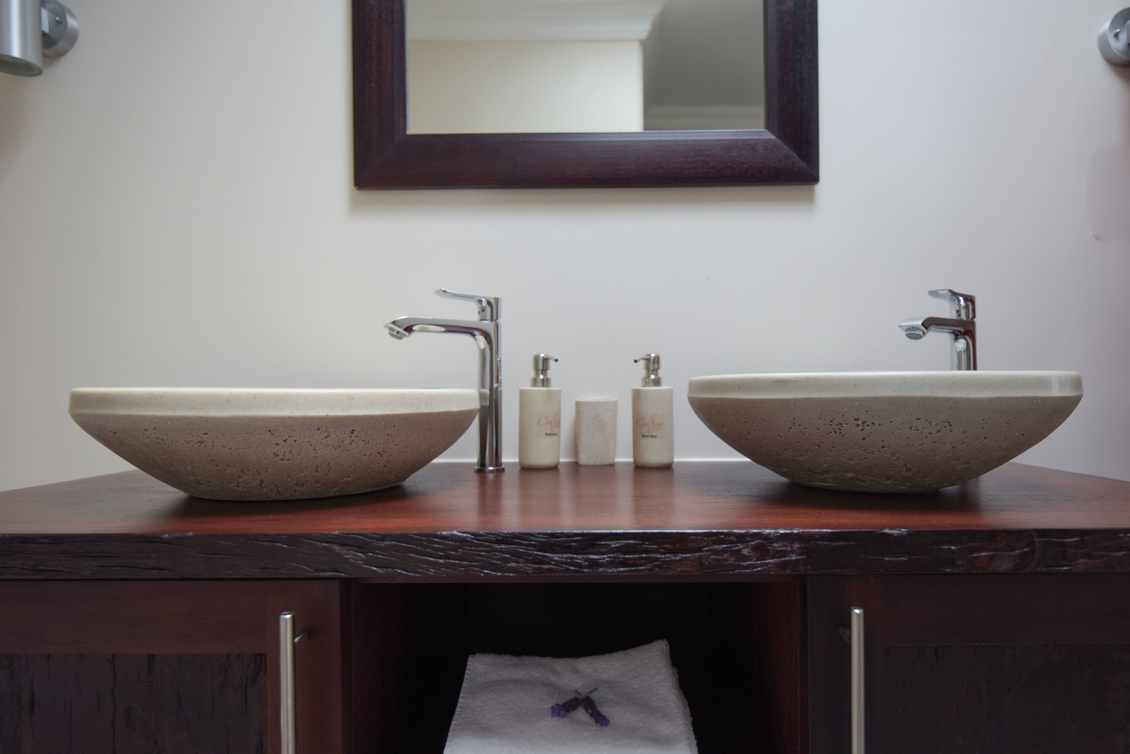 A bathroom with two large, beige, bowl-shaped sinks, a dark wooden countertop, a mirror above the sinks, and a towel hanging in a cabinet below.