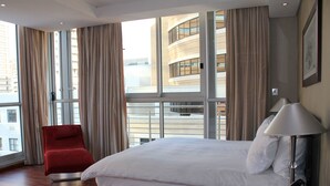 Camera d'albergo con un letto grande, una poltrona rossa, un comodino con una lampada e vista su un edificio attraverso ampie finestre.