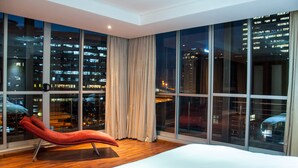 Una camera d'albergo moderna con un letto grande, una poltrona rossa e vista sulla città attraverso ampie finestre.