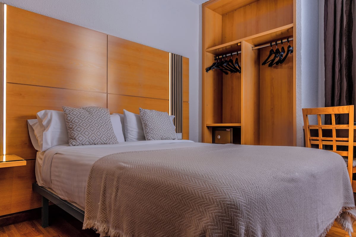 A neatly made bed with patterned pillows, a wooden headboard, and a wooden wardrobe with hanging clothes.
