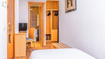 A compact room with a bed, a desk, a chair, a TV, a wooden wardrobe, and a shower area.