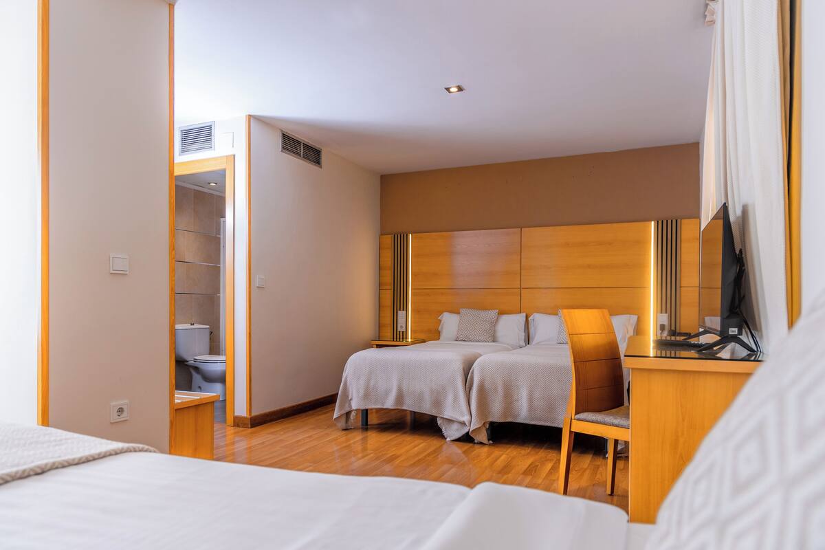 A hotel room with a wooden headboard, two beds, a desk with a computer, and a bathroom visible through an open door.