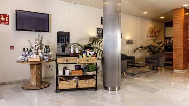 A modern lobby with a marble floor, a round table with various items, a coffee machine, a large pillar, and a TV mounted on the wall.
