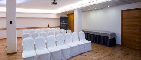 Un salón de conferencias con filas de sillas cubiertas de blanco, un escenario con un atril y un cartel que dice 'SERRANO'.