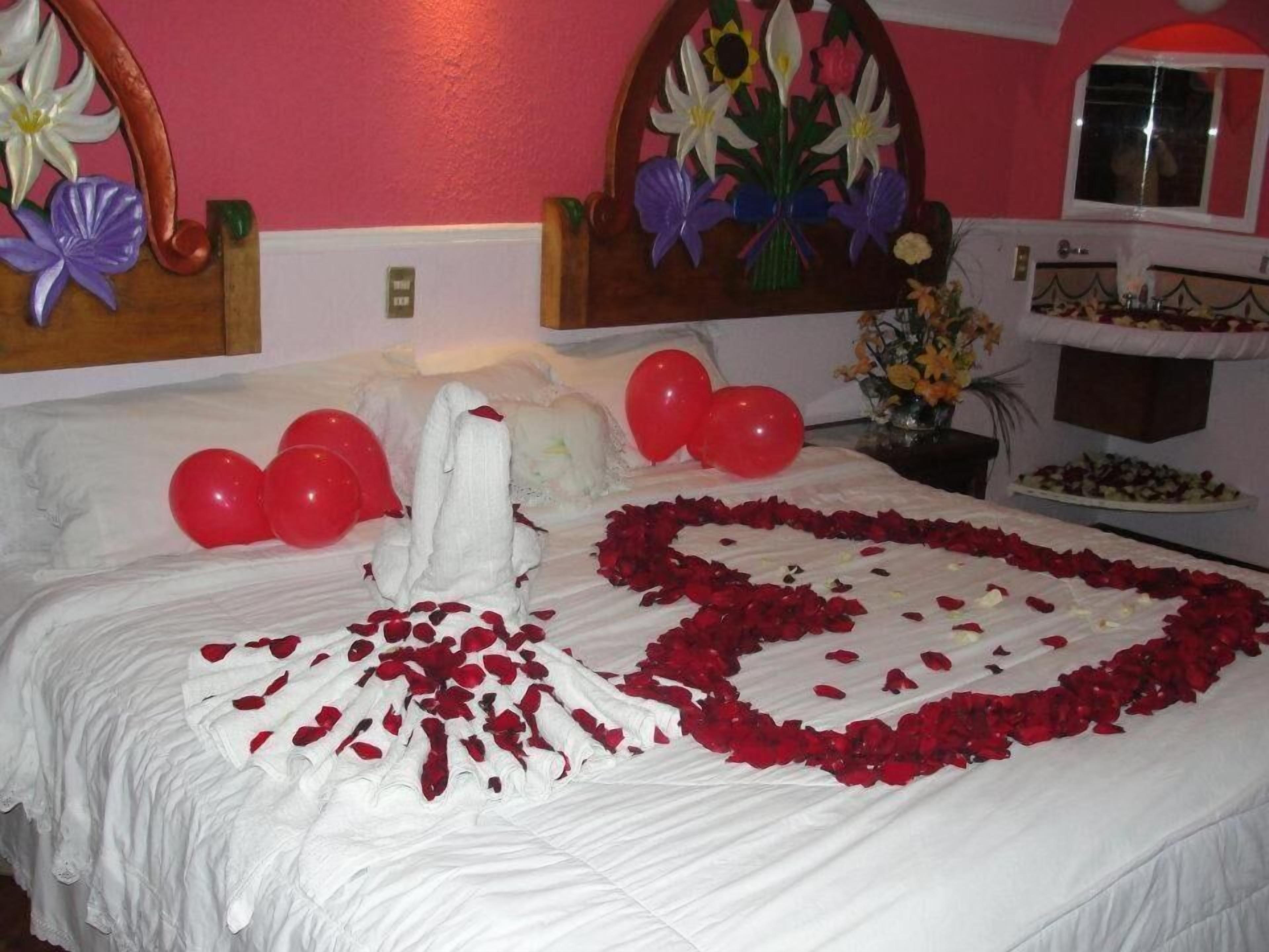 Una habitación de hotel con una cama decorada con pétalos de rosa y globos, un cabecero floral y una pequeña mesa con un jarrón de flores.