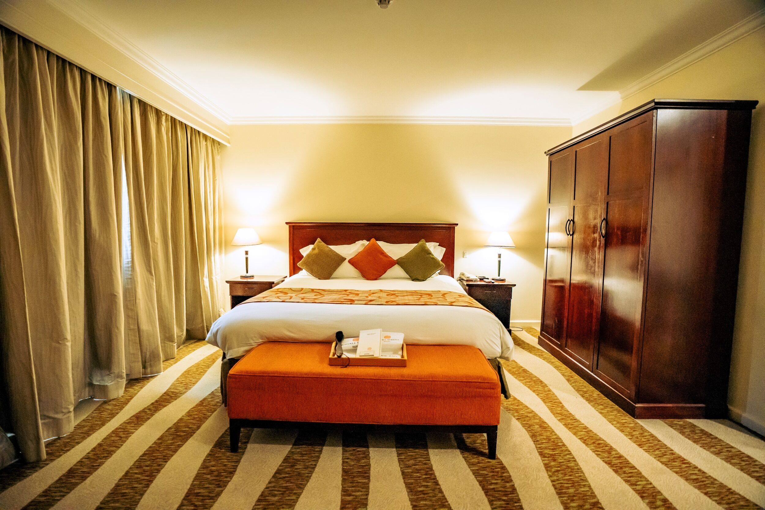 A hotel room with a large bed, two bedside lamps, a wooden wardrobe, and a striped carpet.