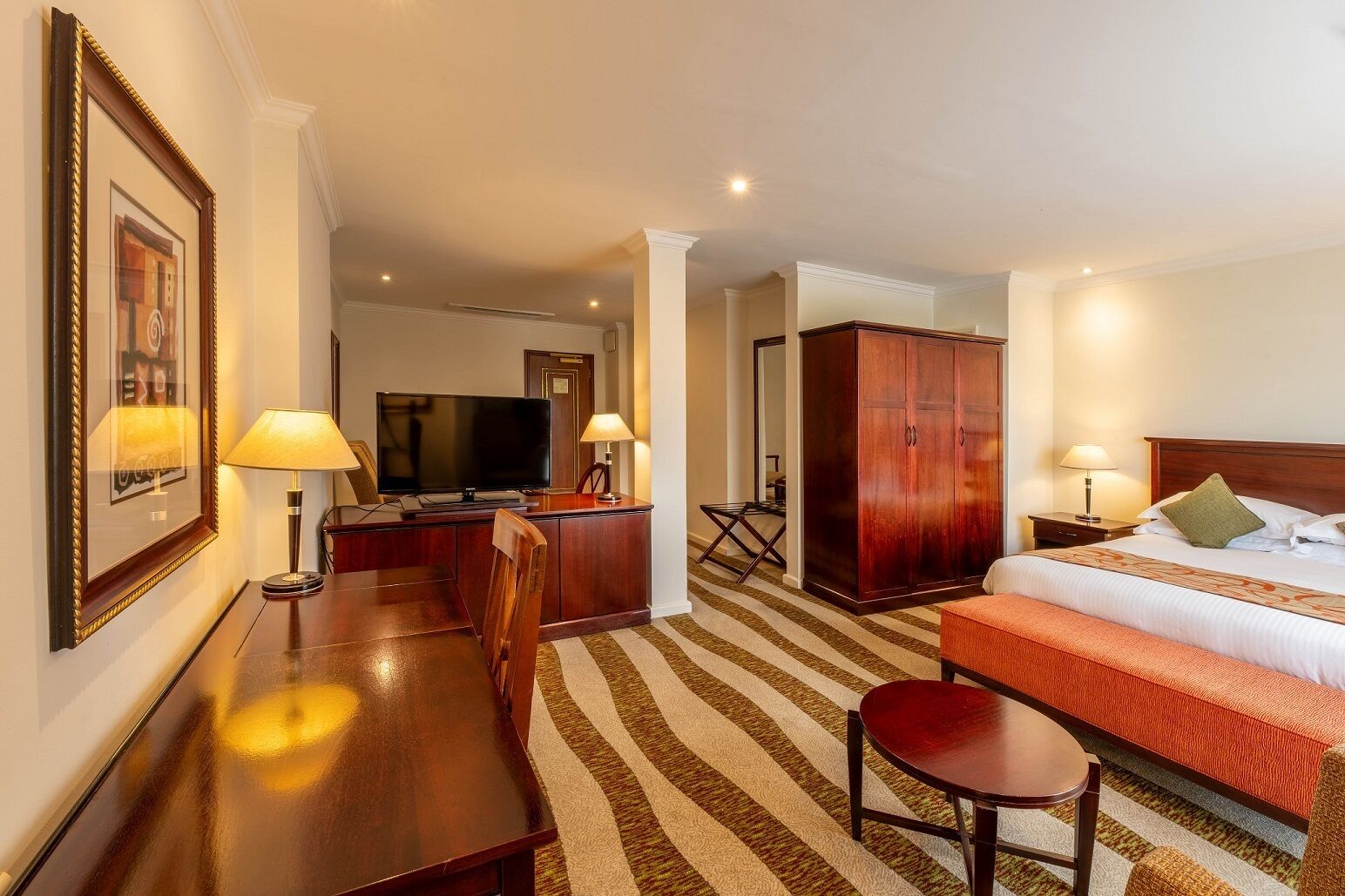 A hotel room with a bed, a desk with a TV, a wardrobe, and a dining area with chairs and a table.