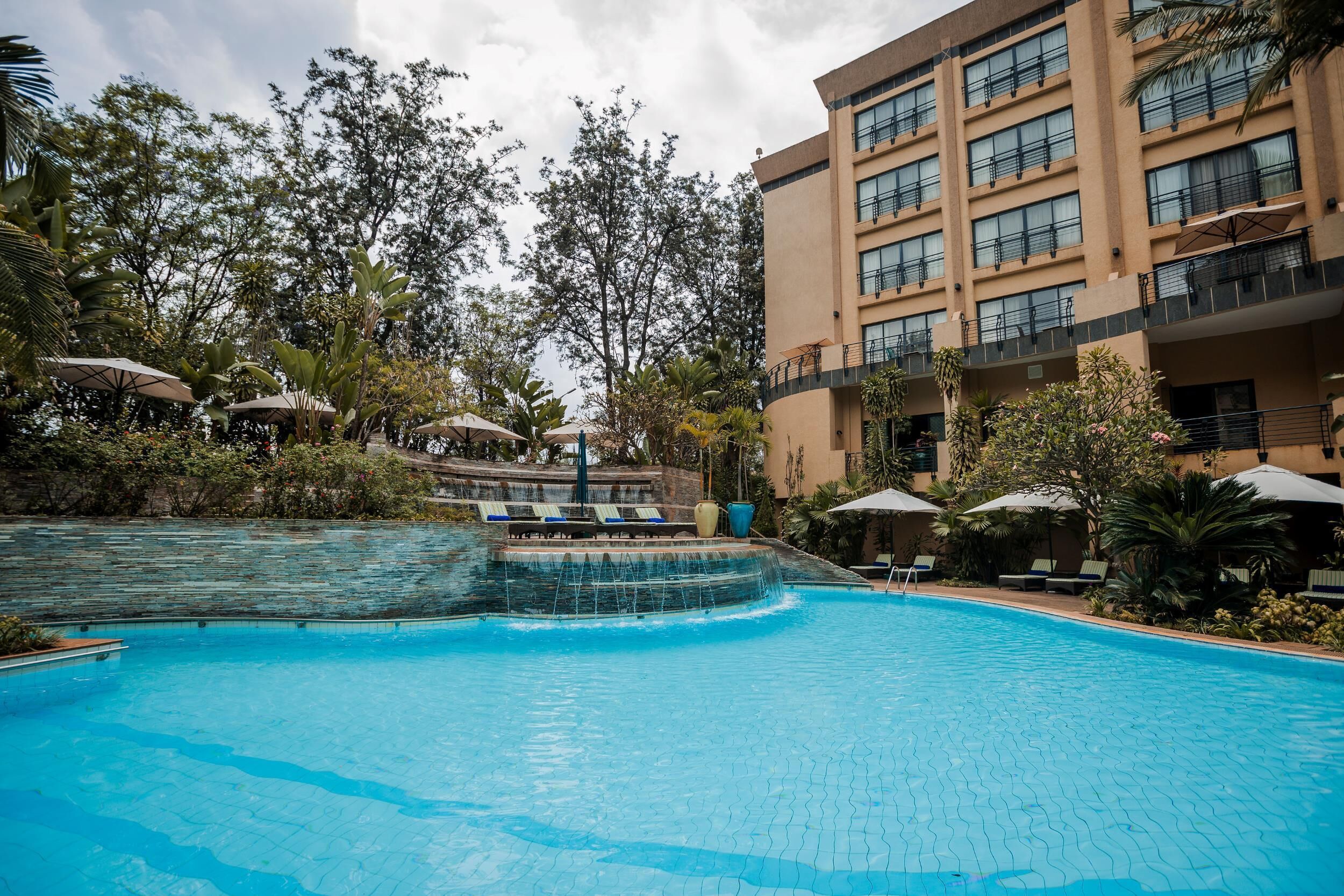 A swimming pool with lounge chairs and umbrellas, surrounded by lush greenery and a multi-story building with balconies.