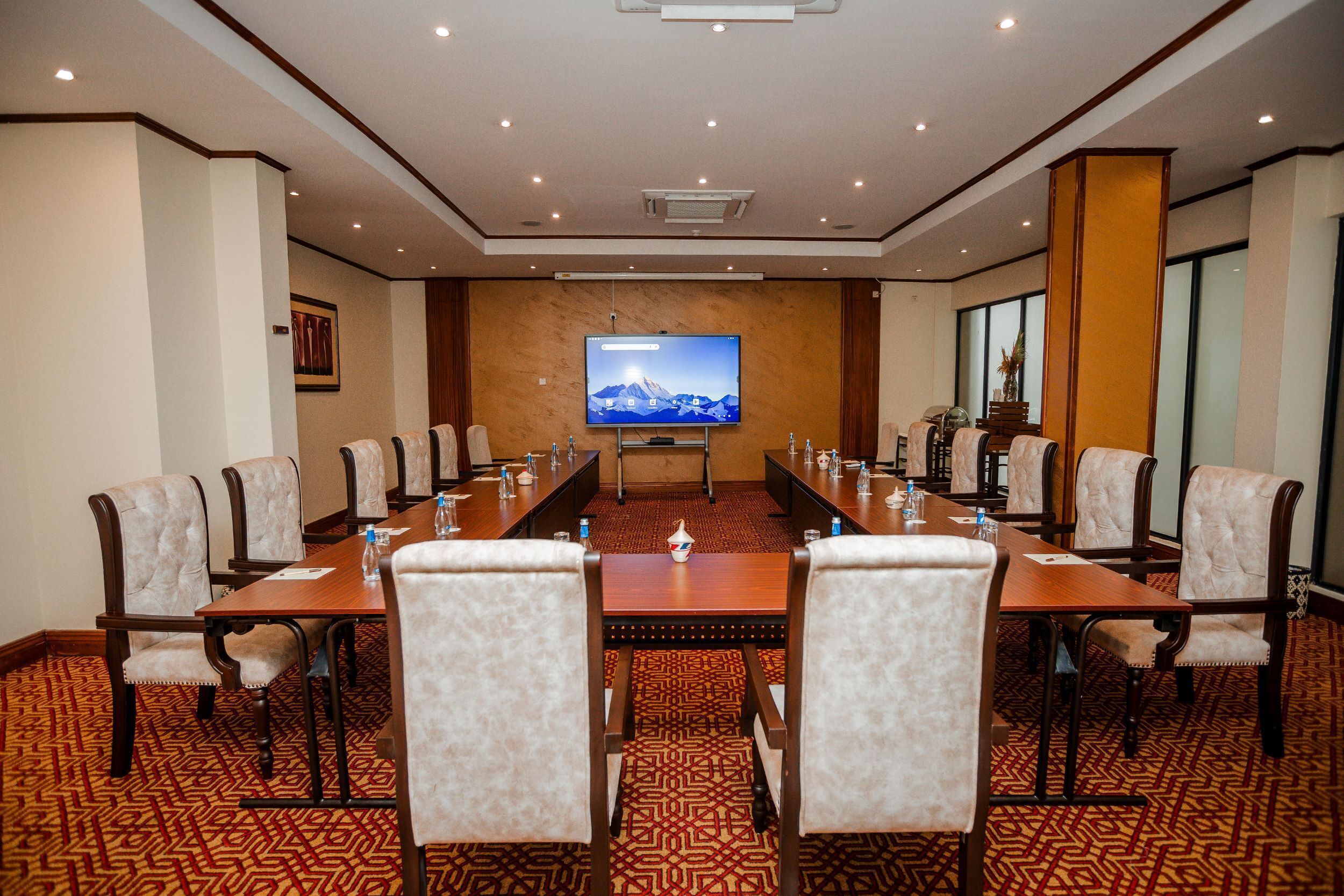 A conference room with a long table, multiple chairs, a television screen, and a patterned carpet.