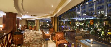A hotel lobby with a patterned carpet, wooden furniture, a glass coffee table, and a view of the outdoor area with umbrellas.