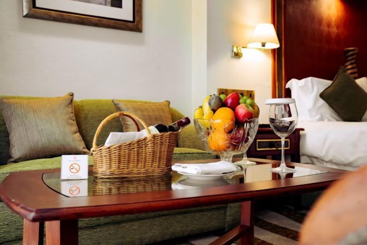 Habitación de hotel con sofá verde, mesa de centro de madera con frutero, copa de vino y una cesta.