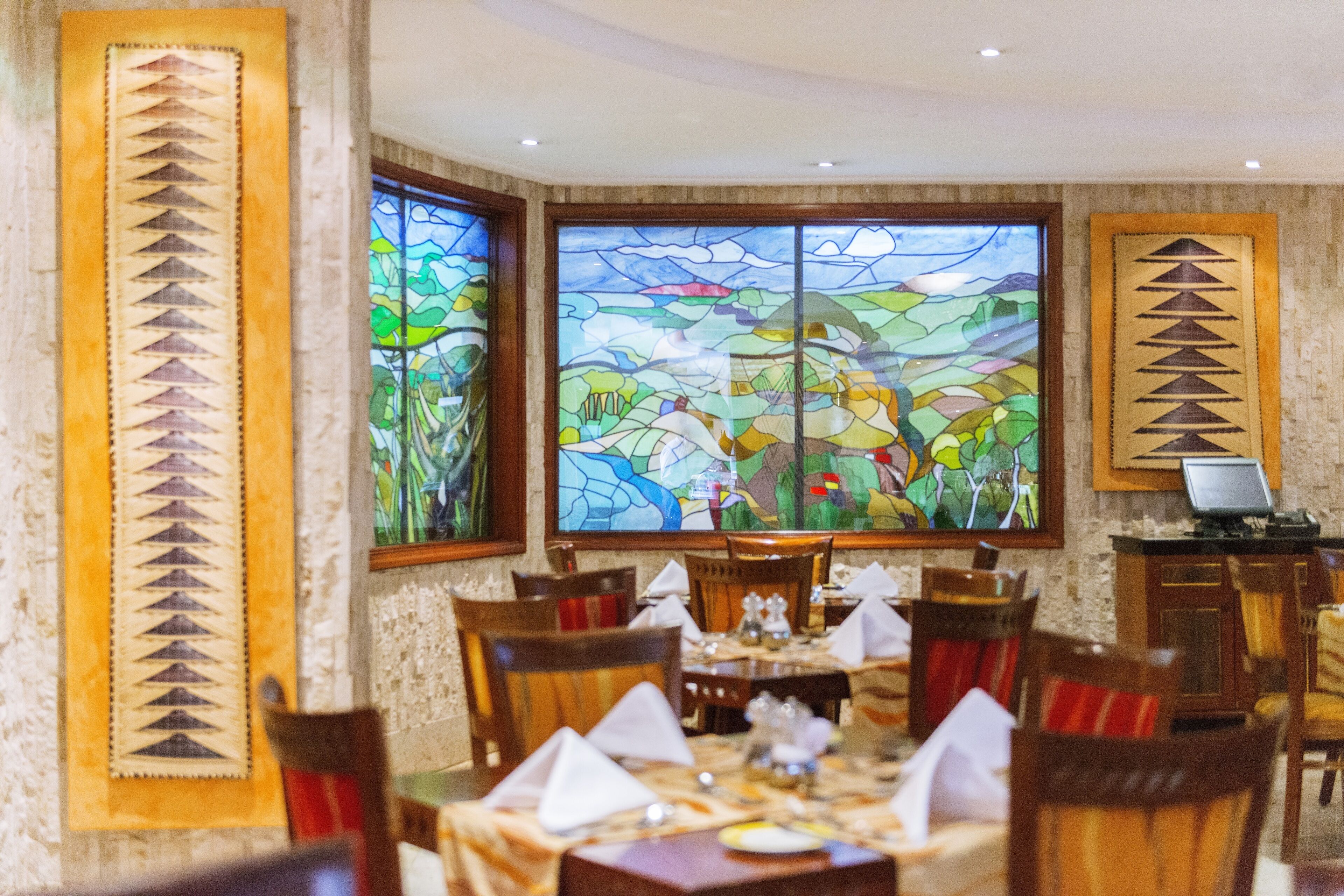 Un comedor con una mesa grande dispuesta para cuatro personas, rodeada de sillas de madera. Hay dos ventanas de vitral grandes con un diseño de paisaje.