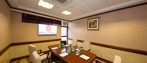 A conference room with a long table, white chairs, and a projector screen displaying the hotel's name.