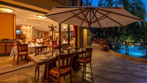 An outdoor dining area with tables and chairs, a large umbrella, and a pool in the background. - Kigali Serena Hotel (Kigali)