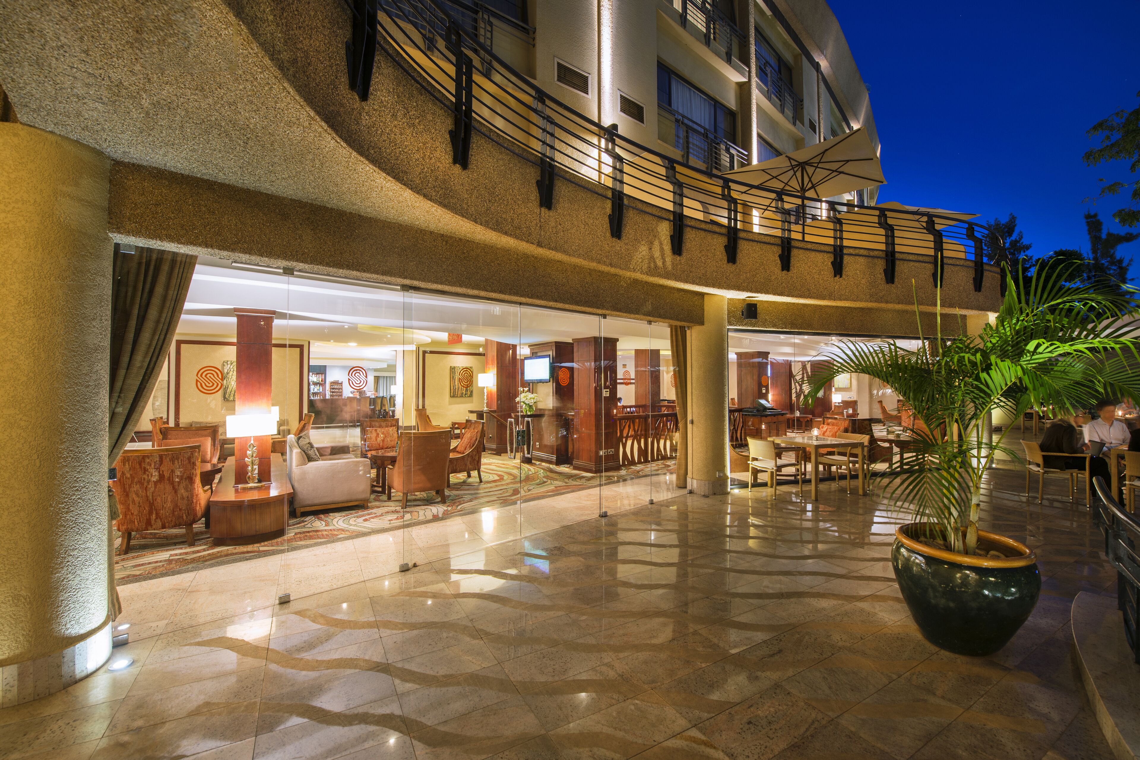 A hotel lobby with a spacious seating area, a large potted plant, and a glass entrance leading to the rooms.