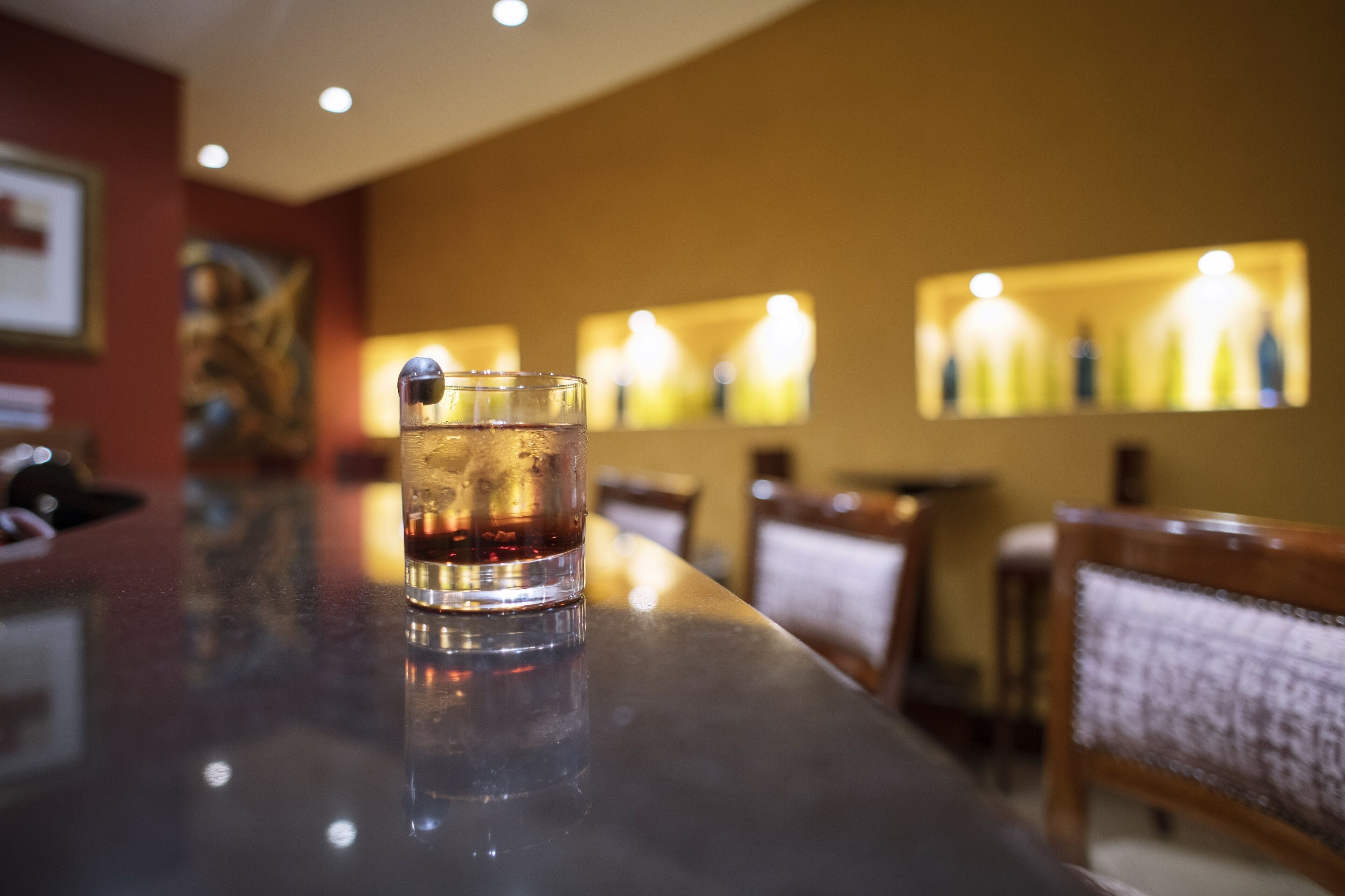 A bar with a glass of dark liquid on the counter, a painting on the wall, and a row of illuminated bottles in the background.