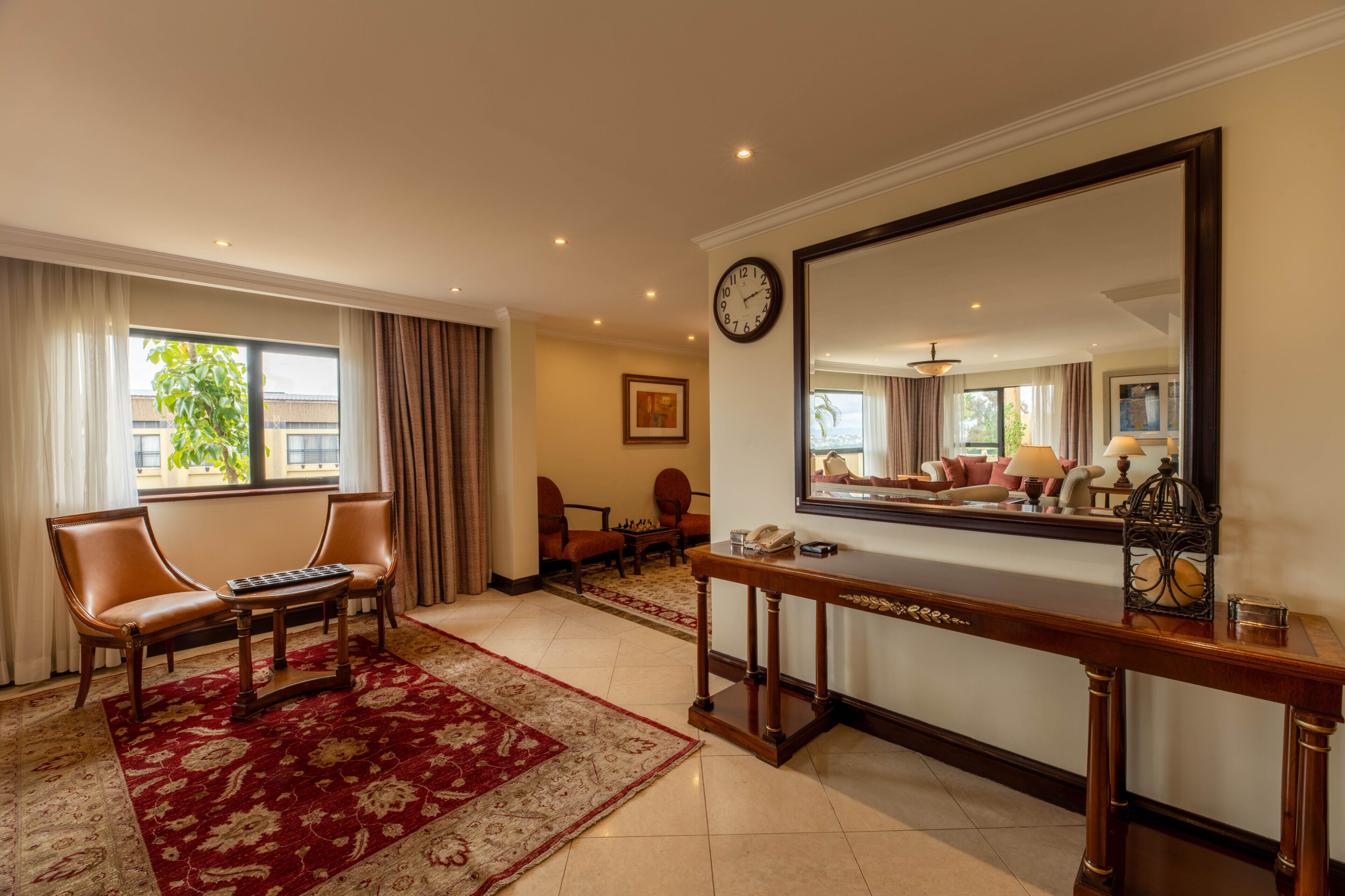 Une chambre d’hôtel avec un grand miroir, un bureau avec un téléphone, un fauteuil en cuir et un tapis rouge à motifs.