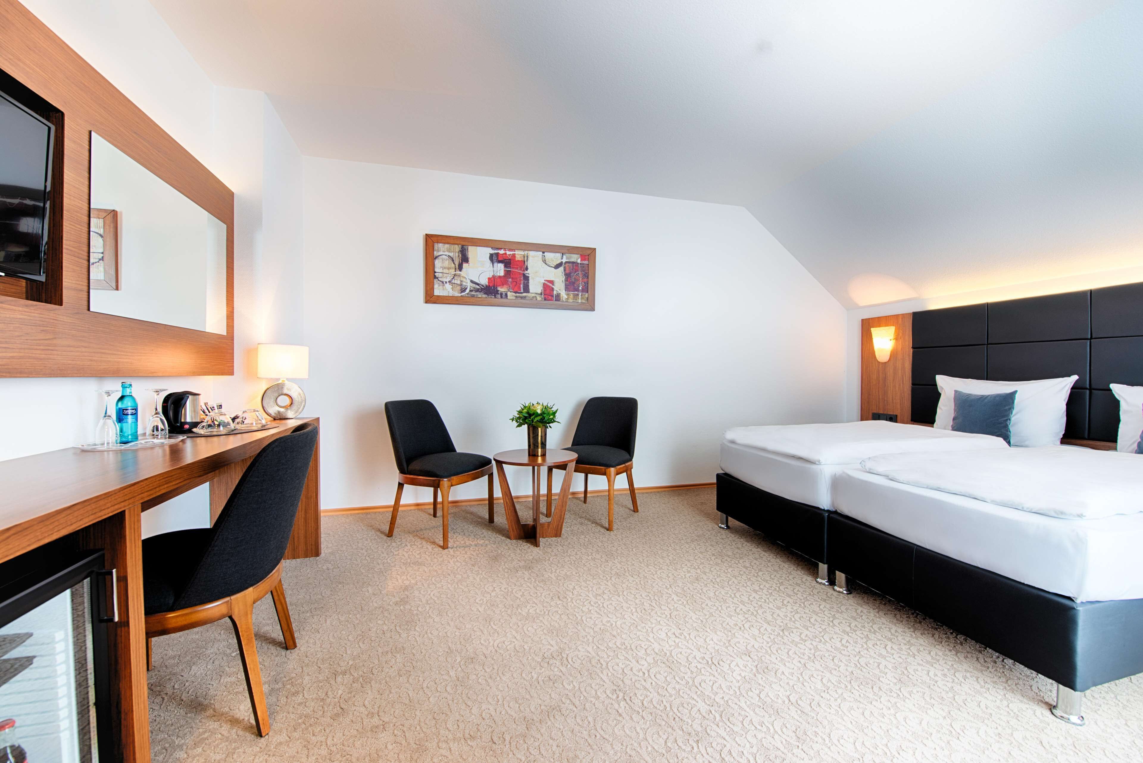 A hotel room with a bed, a desk with chairs, a small table with a plant, and a painting on the wall.