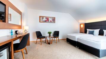 A hotel room with a bed, a desk with chairs, a small table with a plant, and a painting on the wall.