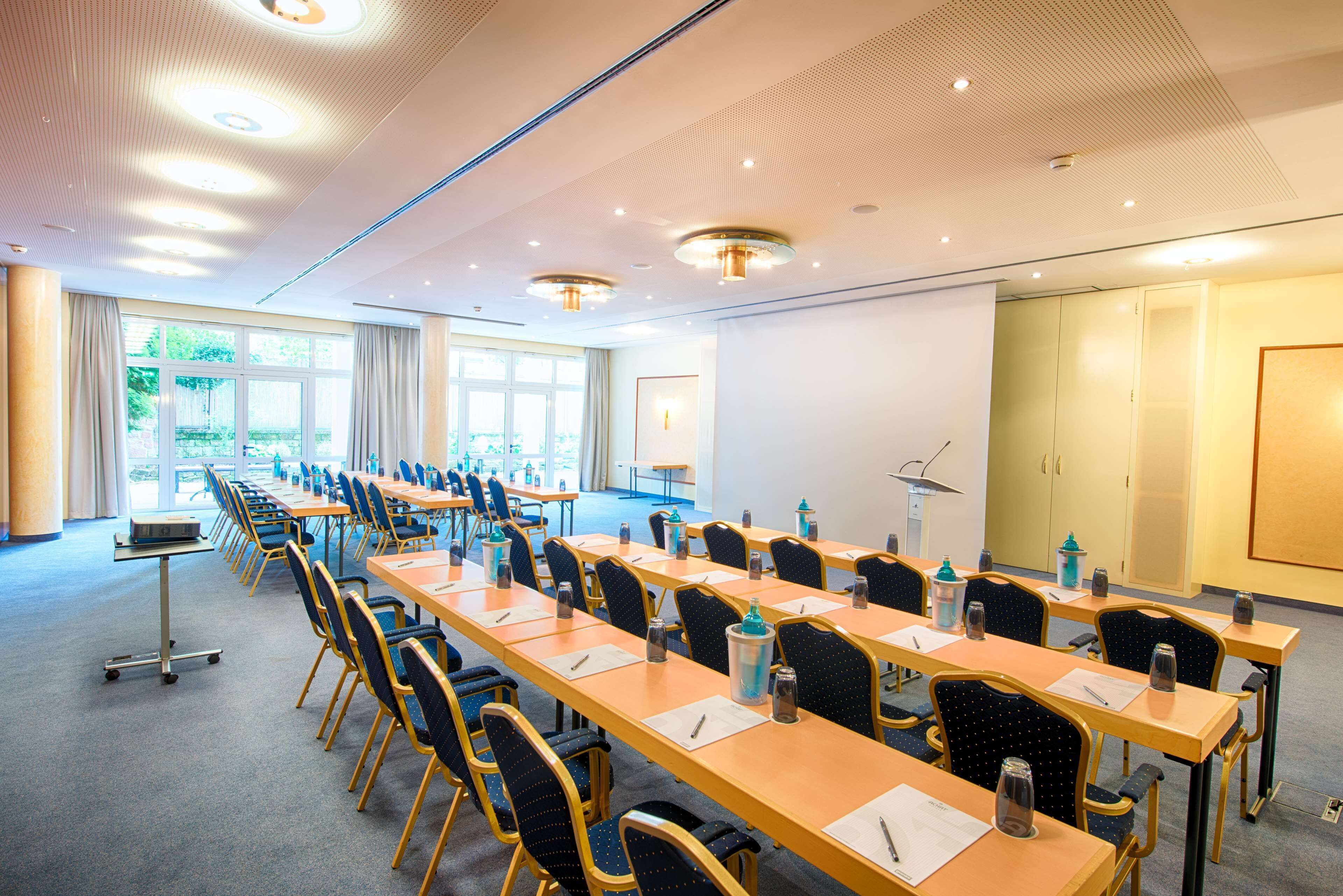Ein Konferenzraum mit langen Tischen und Stühlen, einem Rednerpult und einem großen Fenster mit Blick auf Grünflächen.