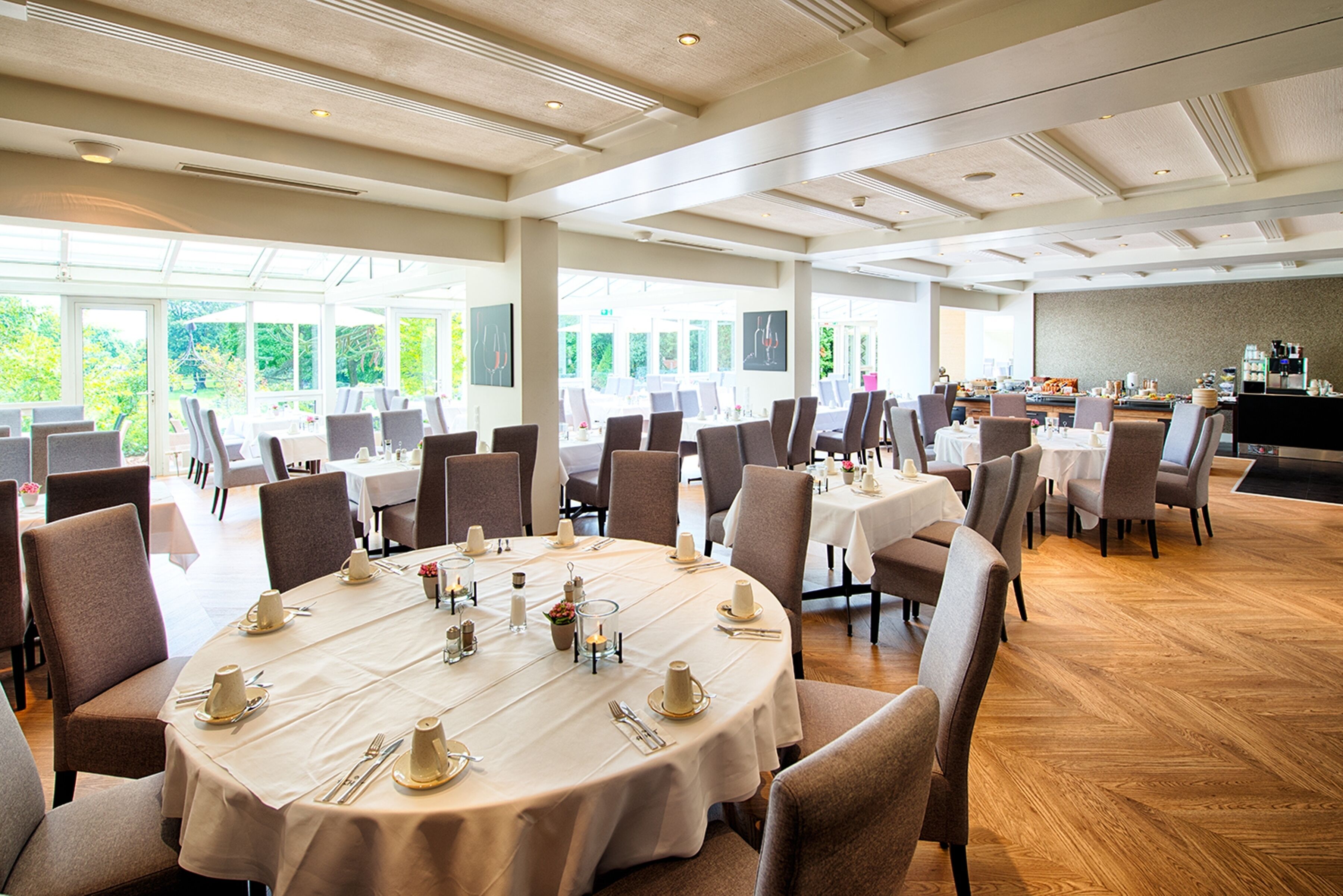 A spacious dining area with round tables set for a meal, large windows offering a view of greenery, and a kitchen area in the background.