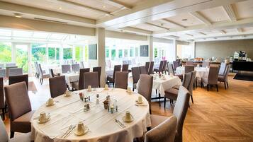 A spacious dining area with round tables set for a meal, large windows offering a view of greenery, and a kitchen area in the background.
