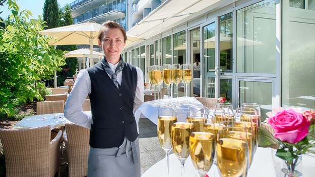 Un serveur se tient près d’une table sur laquelle sont disposés plusieurs verres de champagne. On aperçoit, en arrière-plan, du mobilier d’extérieur en osier et une structure de bâtiment en verre.
