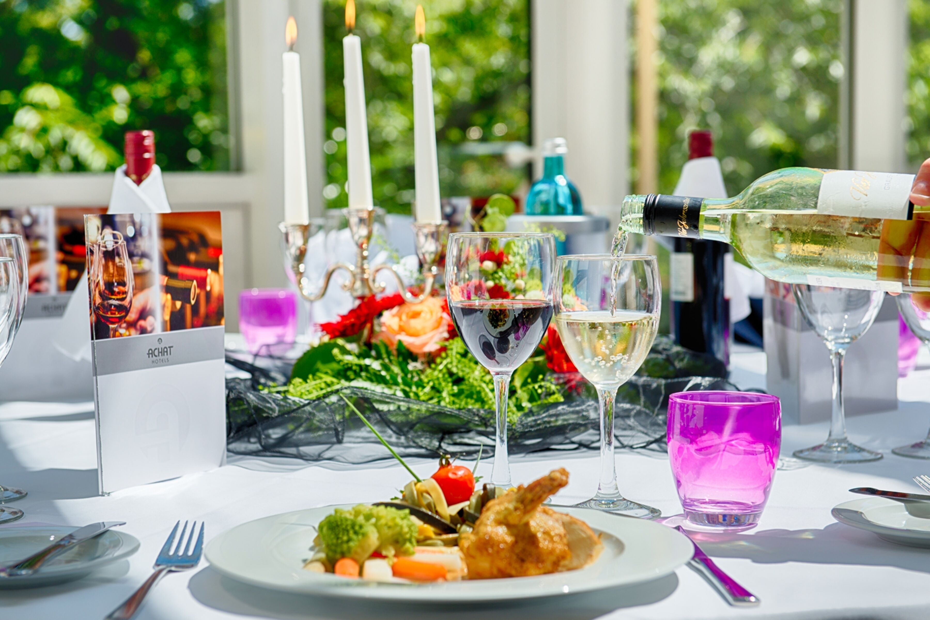 A table set for a meal with a bottle of white wine being poured into a glass, surrounded by plates of food, glasses, and candles.