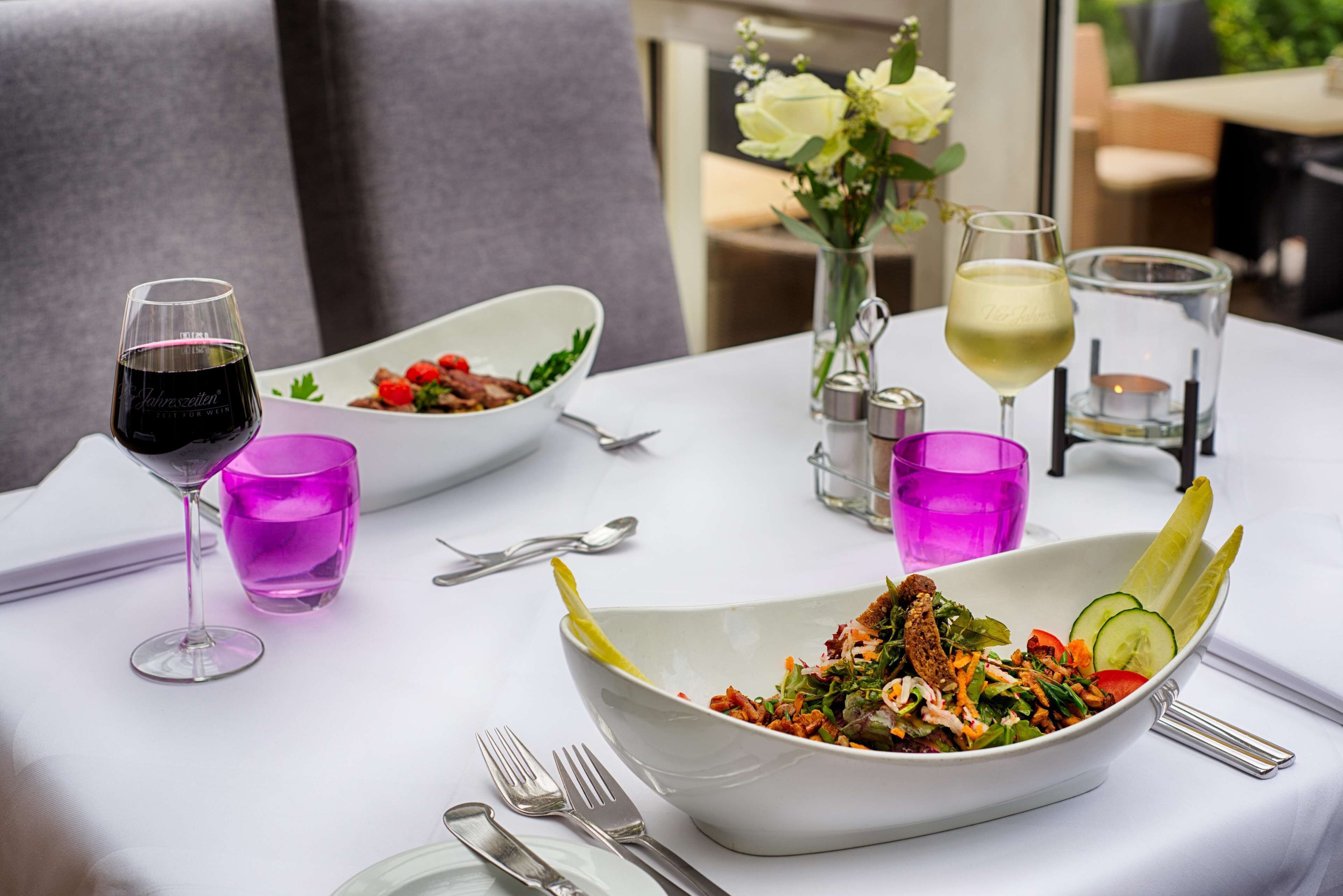 A dining table with a white tablecloth, two white bowls of salad, a glass of red wine, a glass of white wine, and a vase with white flowers.