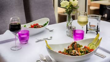 A dining table with a white tablecloth, two white bowls of salad, a glass of red wine, a glass of white wine, and a vase with white flowers.