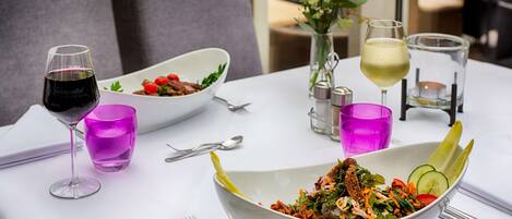 A dining table with a white tablecloth, two white bowls of salad, a glass of red wine, a glass of white wine, and a vase with white flowers.