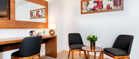 A hotel room with a wooden desk, two chairs, a round table with a plant, a mirror, and abstract artwork on the wall.