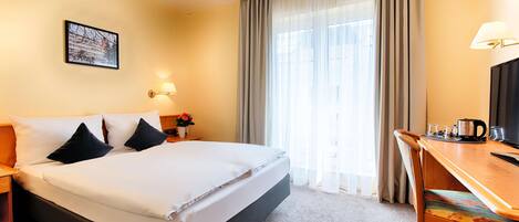 Habitación de hotel con cama, escritorio, silla, lámpara, un cuadro en la pared y ventana con cortinas.
