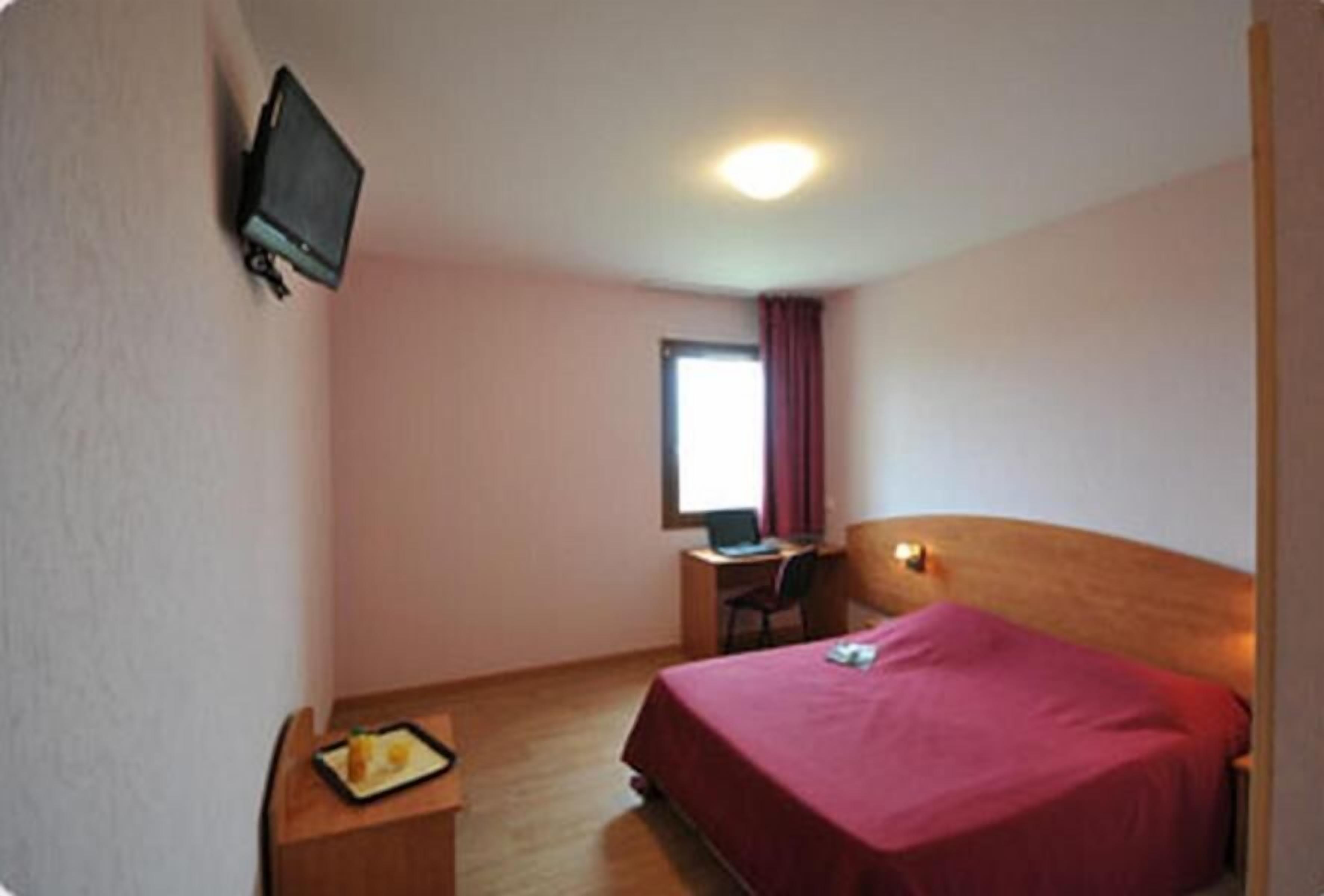 Ein Hotelzimmer mit einem Bett, einem Schreibtisch mit Computer, einem an der Wand montierten Fernseher und einem Fenster mit Vorhängen.