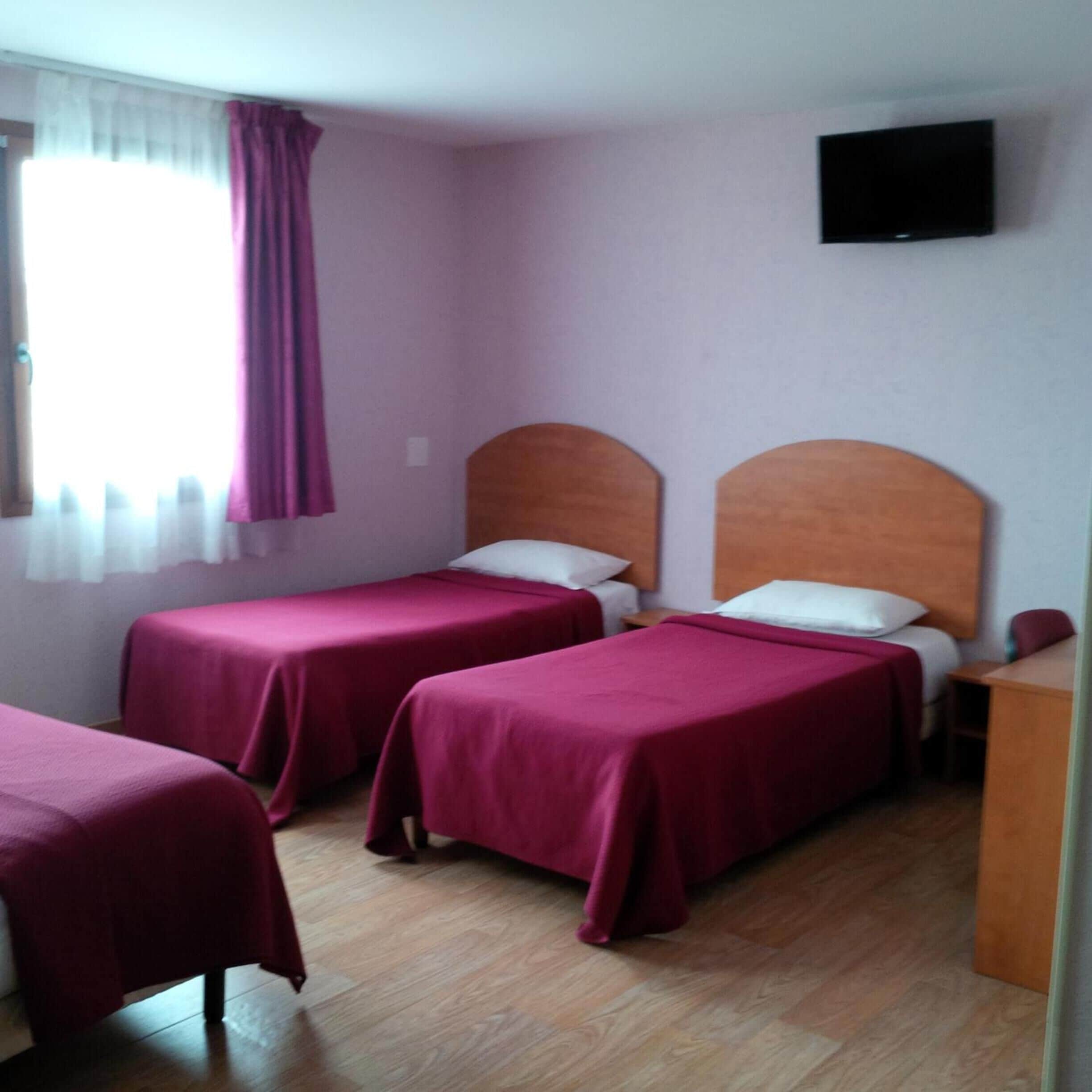 A hotel room with two single beds, a wooden headboard, a television on the wall, and a window with curtains.