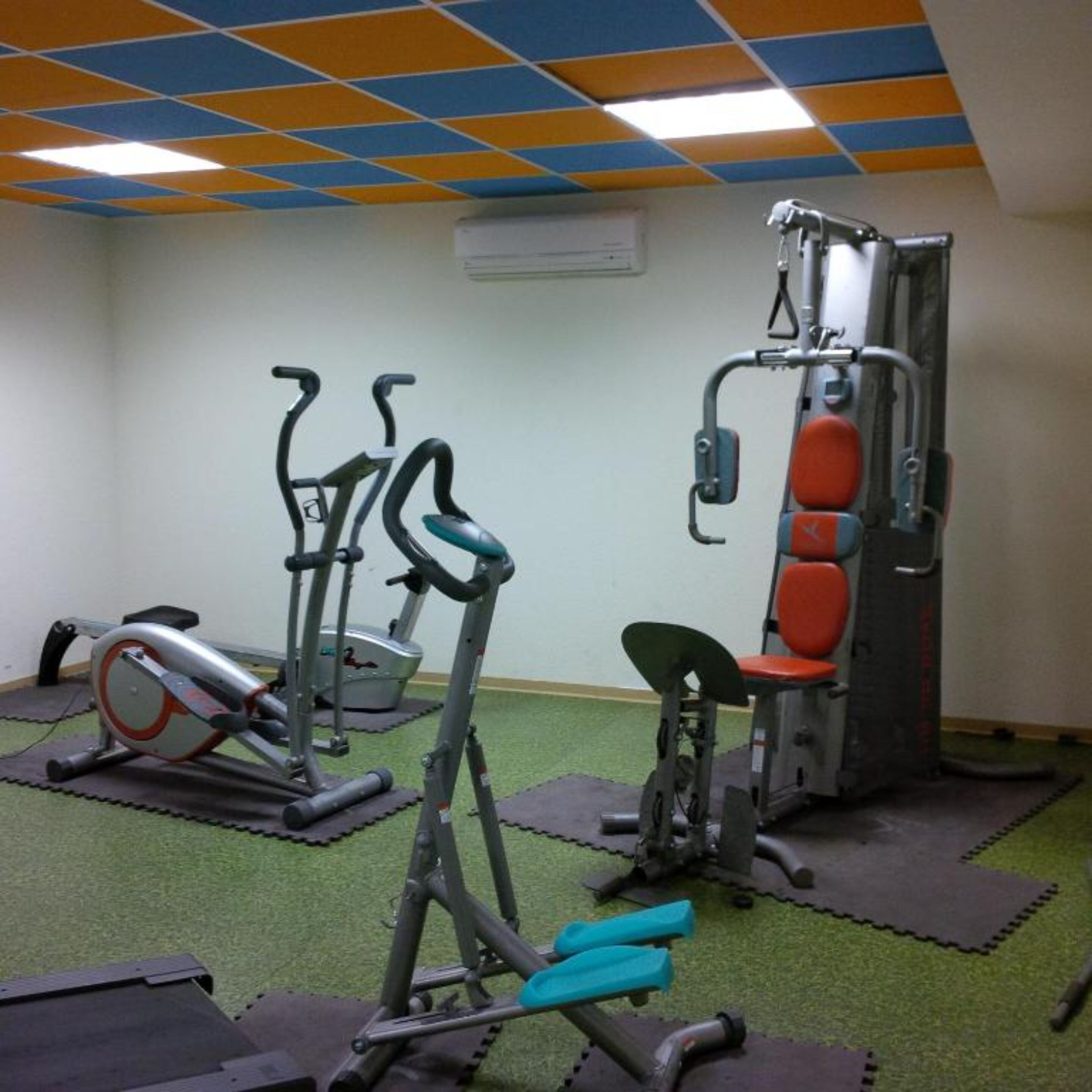 Un gimnasio con equipo de ejercicio, incluyendo cintas de correr y una máquina de pesas, sobre colchonetas verdes.
