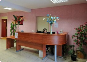 Un mostrador de recepción con detalles de madera, dos plantas en macetas y un suelo de baldosas.