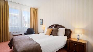 Quarto de hotel com uma cama grande, uma mesa de cabeceira com abajur, uma cadeira e vista da cidade através de cortinas translúcidas.