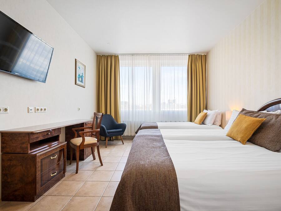 A hotel room with a flat-screen TV, a desk with drawers, a chair, two beds with pillows, and a large window with curtains.