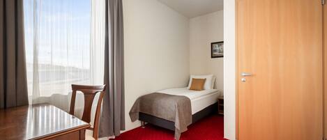 Ein Hotelzimmer mit einem Bett, einem Holztisch, einem Stuhl, einem roten Teppich und einer Holztür.