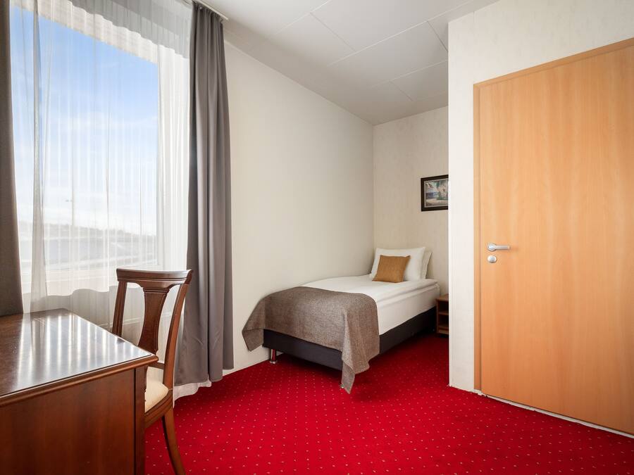 Een hotelkamer met een bed, een houten bureau, een stoel, een rood tapijt en een houten deur.