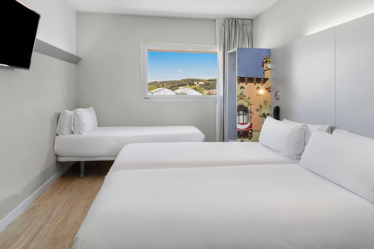 A hotel room with two beds, a window with a view of buildings, a flat-screen TV mounted on the wall, and a decorative wall piece.