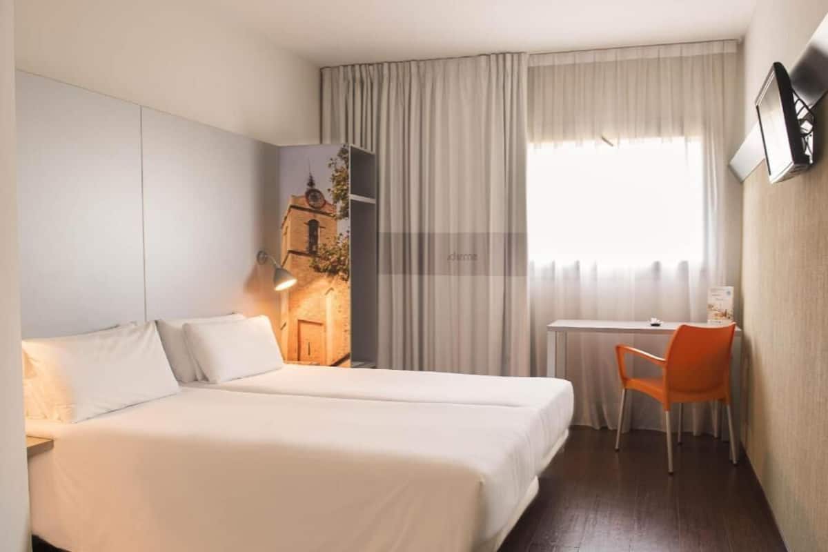 Una habitación de hotel con dos camas, un escritorio, una silla, una lámpara y una ventana con cortinas.